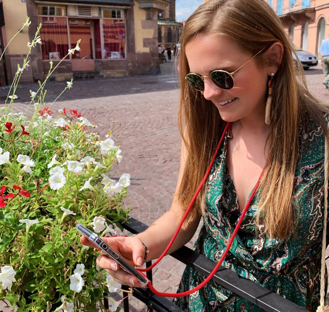 Frau fotografiert mit Handy, dass sie an roter Handykette am Hals umgehangen hat.
